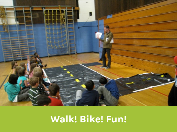 Image of a "Walk bike fun" activity in a school gym. An instructor is standing up and teaching to children sitting on the ground.
