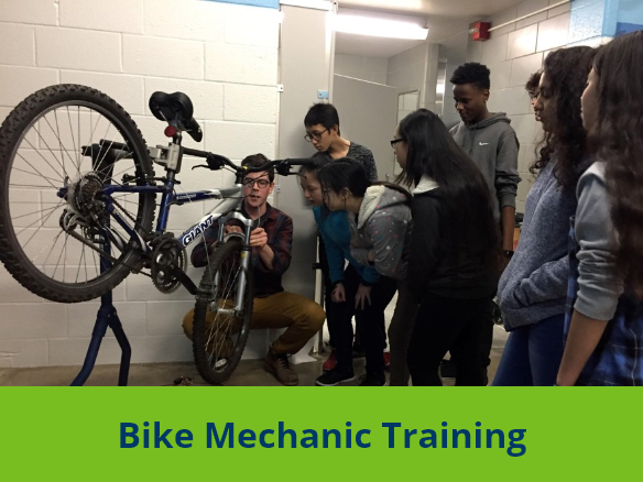 Image of a bike mechanic knelt in front of a bike propped on a stand, with students gathered around for a demonstration.